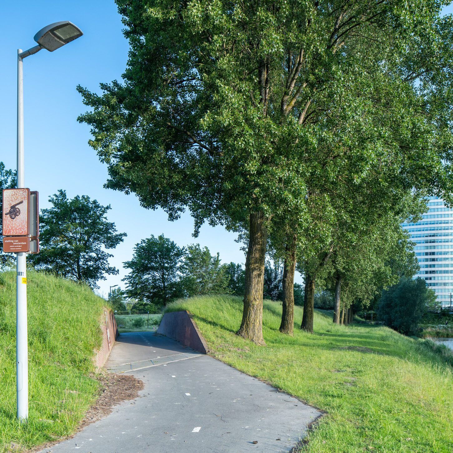 Beeldpaneel Kanon langs de Geniedijk met doorgang door de dijk en moderne kantoorgebouwen op de achtergrond