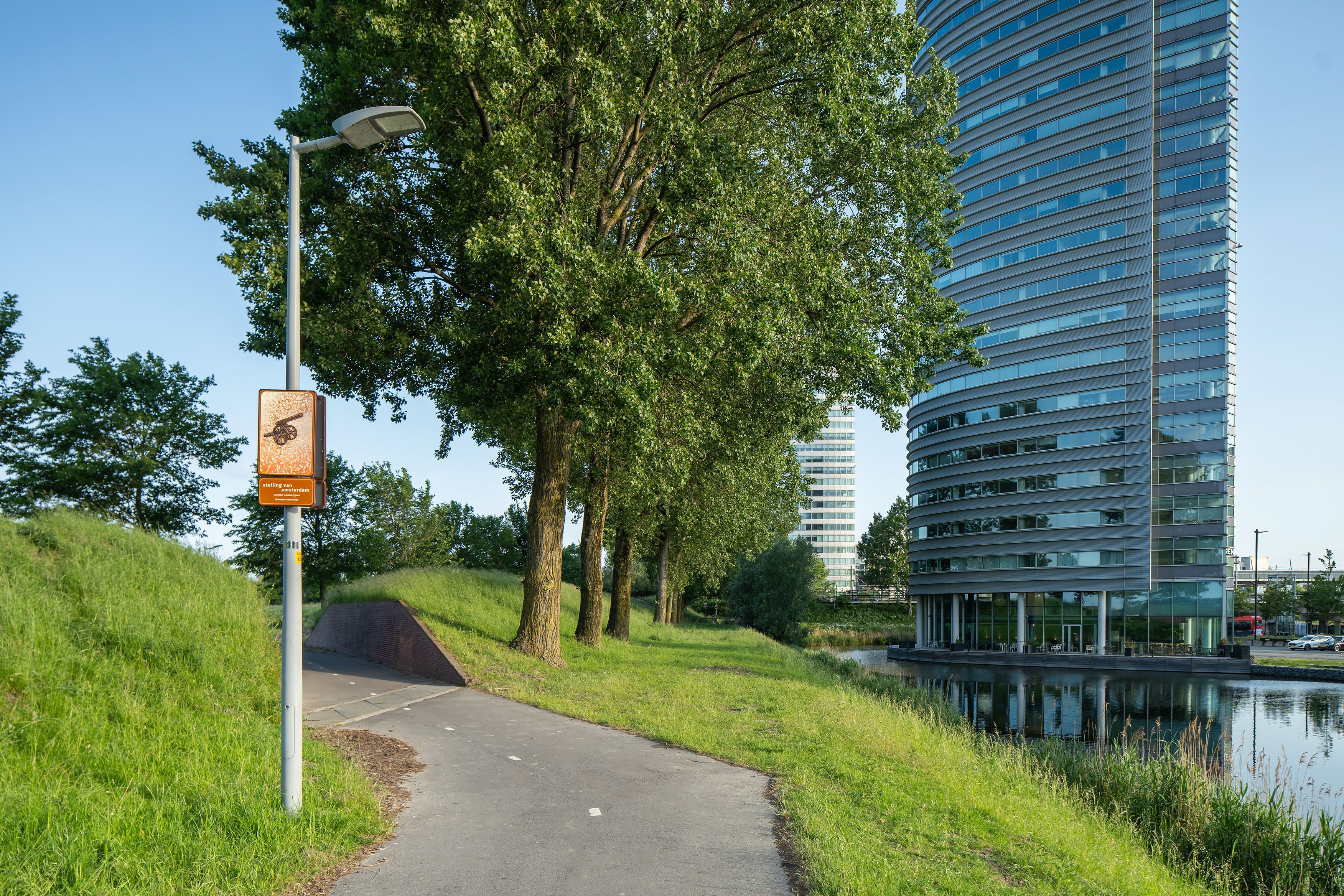 Beeldpaneel Kanon langs de Geniedijk met doorgang door de dijk, watergang en moderne bebouwing van Hoofddorp